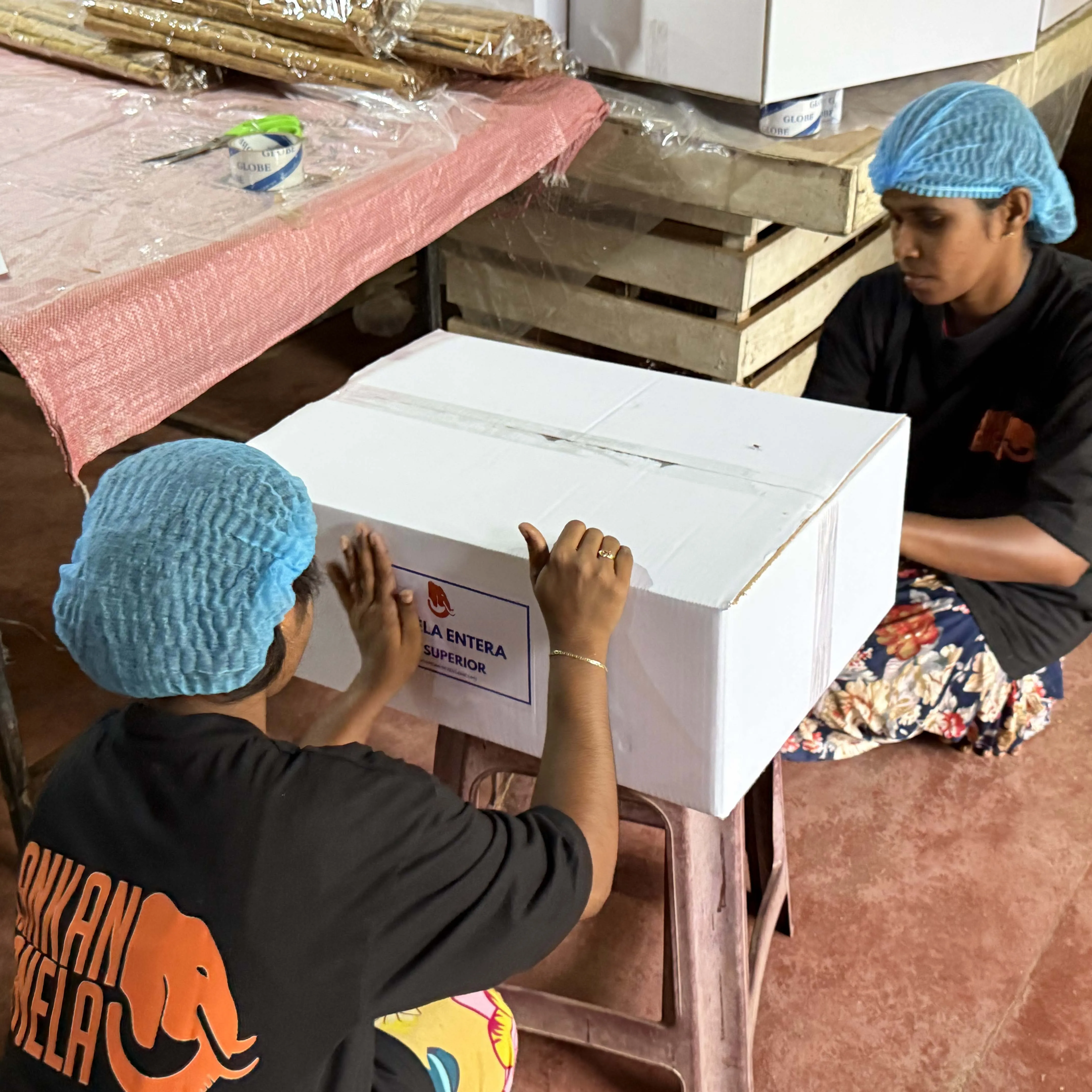 Two women wearing blue hairnets and black t-shirts with the "Lankan Canela" logo are preparing a shipment. The woman on the left is carefully applying a label that reads "LA ENTERA SUPERIOR" to a white cardboard box, while the woman on the right sits opposite her. They are in a workshop setting with a table holding cinnamon quills and tape, and stacked wooden crates in the background.