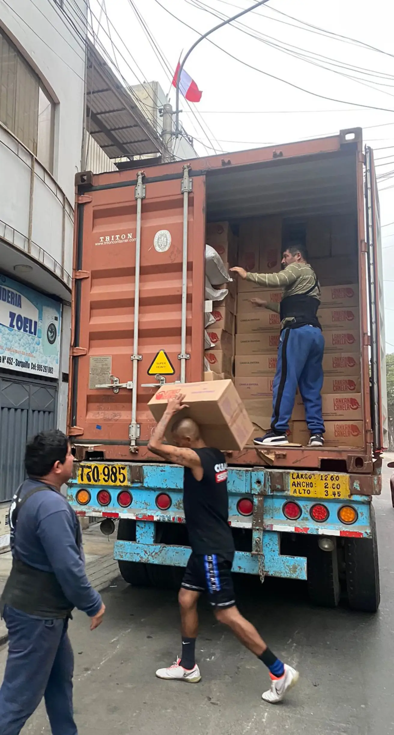 Workers unload a shipping container filled with Lankan Canela branded boxes on a city street. One man carries a large carton on his shoulder, while another organizes the stacks inside the container. A Peruvian flag flies above the scene, indicating the arrival of the cinnamon shipment at its international destination in Peru.